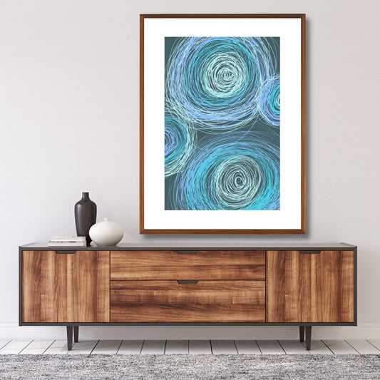 Grafisk plakat med dekorativt sirkelmotiv i friske blå og grønne nyanser på mørk blågrønn bunn. Plakaten har en hvit kant som gir mer dybde og fremhever motivet. Illustrasjonen viser plakaten i eikeramme, som henger på en vegg over en skjenk.