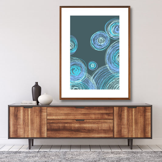 Grafisk plakat med dekorativt sirkelmotiv i friske blå og grønne nyanser på mørk blågrønn bunn. Plakaten har en hvit kant som gir mer dybde og fremhever motivet. Illustrasjonen viser plakaten i eikeramme, som henger på en vegg over en skjenk.