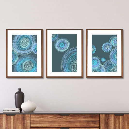 En serie plakater med dekorative, grafiske sirkelmotiv i friske blå og grønne nyanser på mørk blågrønn bunn. Plakatene har en hvit kant som gir mer dybde og fremhever motivet. Illustrasjonen viser plakatene i eikerammer, som henger på en vegg over en skjenk.