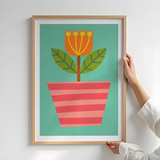 Bold Botanical No13 Er en oransje blomst med gule støvbærere strekker seg opp fra en rød- og rosestripet potte. To grønne blader med mørke årenett utfolder seg mot en turkis bakgrunn, og gir motivet et friskt og energisk uttrykk. Illustrasjon viser plakat i eik ramme.