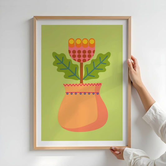 Bold Botanical No14 med sinn fantasifulle blomst med oransje og rosa kronblader prydet med røde prikker reiser seg fra en rund, dekorert vase i varme ferskentoner. To taggete blader i grønt rammer inn motivet, og skaper en leken kontrast mot den lysegrønne bakgrunnen. Illustrasjon viser plakat i eik ramme.