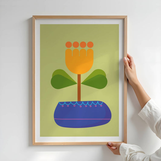 Bold Botanical No16 viser et stilisert blomsterhode i glødende oransje og gult vokser opp fra en dypblå potte med rosa og turkise detaljer. To bladpar i grønn gradient gir et harmonisk balansepunkt mot den lys olivengrønne bakgrunnen, og gir motivet et moderne og leken uttrykk. Illustrasjon viser plakat i eik ramme.