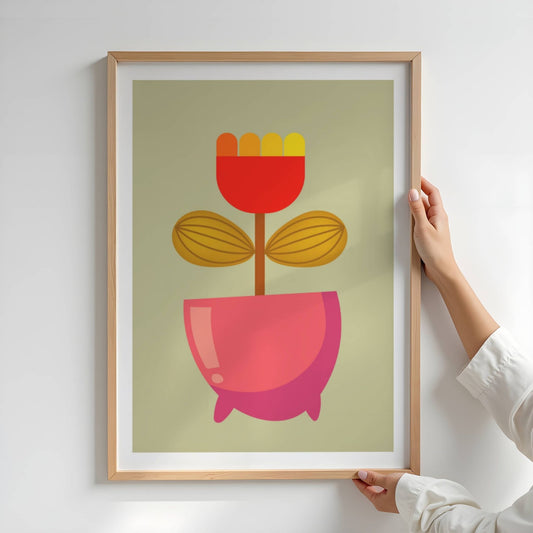 Bold Botanical No17 viser et kompakt blomsterhode i rød bunnfarge med varme nyanser fra oransje til gult kronblad gir et kraftfullt inntrykk. De ovale, stripete bladene i gyllenbrun farge balanserer det sterke uttrykket, mens en rødrosa blomsterpotte med myk glans gir motivet en leken og moderne karakter. Illustrasjon viser plakat i eik ramme.