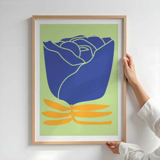 Bold Botanical No3 er et grafisk blomstermotiv med en dyp kongeblå rose i full knopp, fremhevet med lyse konturlinjer. Den står stødig på et sett stiliserte, oransje bladformer som gir motivet en balansert og moderne kontrast mot den sarte, grønne bakgrunnen. Illustrasjon viser plakat i eik ramme.