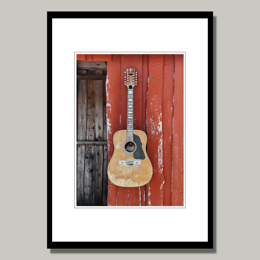 Motivet av en slitt og velbrukt kassegitar som henger på det rødmalte buss-stoppet på Toftarøyna fanger essensen av norsk kystkultur på en vakker og nostalgisk måte. Illustrasjonsfoto viser motivet som plakat i svart ramme.