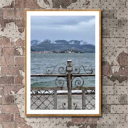 En regntung og vindfull augustdag i Stresa ved Lago Magiore, Piemonte. Det er lavt skydekke og bølger på sjøen. Et gjerde i smijern skiller parken og sjøen. I bakkant skimtes Alpene og byen Verbania.