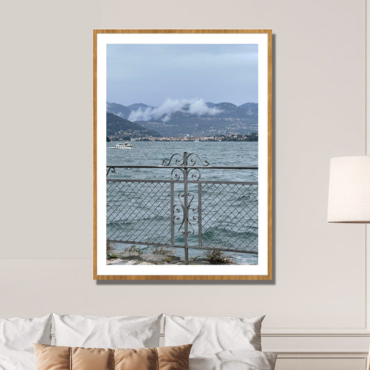 En regntung og vindfull augustdag i Stresa ved Lago Magiore, Piemonte. Det er lavt skydekke og bølger på sjøen. Et gjerde i smijern skiller parken og sjøen. I bakkant skimtes Alpene og byen Verbania.