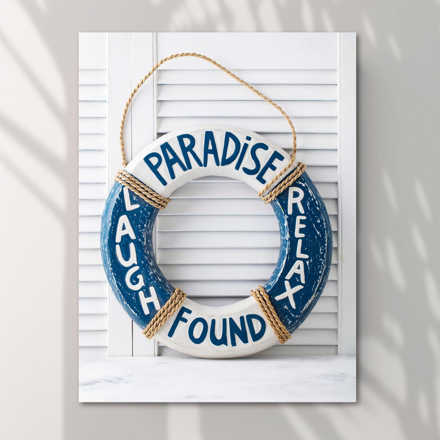 Motivet viser en dekorativ livbøye i elegante blå og hvite nyanser, som står foran en klassisk hvit sjalusidør. Den er påført de inspirerende ordene PARADISE, LAUGH, FOUND og RELAX. Illustrasjonsfoto viser motivet på lerret.
