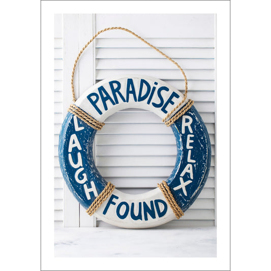Motivet viser en dekorativ livbøye i elegante blå og hvite nyanser, som står foran en klassisk hvit sjalusidør. Den er påført de inspirerende ordene PARADISE, LAUGH, FOUND og RELAX.