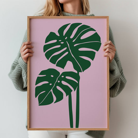 Monstera Blush – en minimalistisk og trendy botanisk plakat med to ikoniske monstera-blader i dyp grønn mot en myk rosa bakgrunn. Illustrasjon viser plakat i eik ramme.