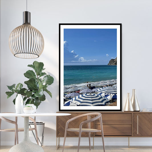 Motiv fra Montereosso al Mare, en av de fem byene i Cinque Terre ved Liguriakysten i Italia. Bildet viser blått middelhav og himmel som helt ned i vannet i horisonten. På høyre side skimtes noen klipper. På sranden er det tett i tett med blå og hvitstripete parasoller. Bildet vise miljøbilde av plakat i sort ramme.