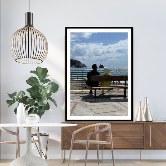 Motiv fra Montereosso al Mare, en av de fem byene i Cinque Terre ved Liguriakysten i Italia. Bildet viser blått middelhav og himmel som helt ned i vannet i horisonten. Her har et par funnet seg godt til rette på en av byens benker. Fri sikt utover Middelhavet så langt øyet ser. Til venstre på bildet går klipper ned i havet. Blå himmel er dekket med noen hvite skyer. Hav og himmel møtes ute i horisonten. Plakaten har en hvit kant rundt og er rammet inn i sort ramme. Rammen står på en benk i et miljø.