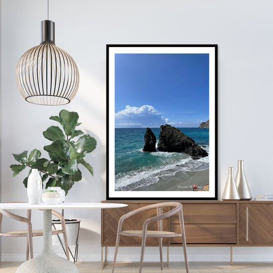 Motiv fra stranden ved Montereosso al Mare, en av de fem byene i Cinque Terre ved Liguriakysten i Italia. Bildet viser blått middelhav og himmel som møter havet i horisonten. . I overgangen mellom himmel og hav ligger en stor skybanke. Det slår små bølger inn mot den karakteristiske klippen som ligger midt på stranden . På bildet er plakaten, som har en hvir kant rundt, satt inn i sort ramme. Den står oppe på en benk i et miljø.