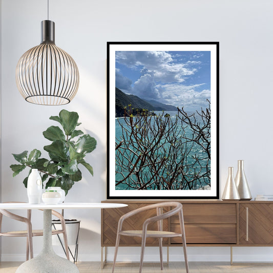 Bildet viser klippene og fjellsiden som går rett ned i havet. Himelen er dekortert med noen skybanker. Havet møter himmelen ute i horisonten. I forgrunnen er det en busk uten blader. Bildet har en hvit kant rundt, og er rammet inn i sort ramme.