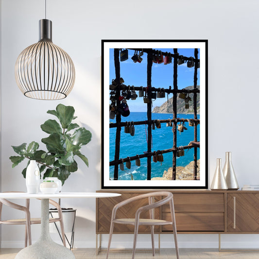 Bildet viser en mur med en åpning med et gitter, der det er hengt opp hengelås med håp om et langt liv i kjærlighet. Gjennom gitteret ses det blåe Middelhavet og klippene som går ned i sjøen. Plakaten har en hvit kant og er rammet inn i sort ramme.