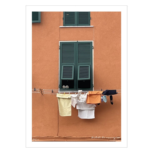 Plakat viser motiv fra Riomaggiore, en av de fem byene i Cinque Terre ved Liguriakysten i Italia. Riomaggiore ligger lengst østover av de 5 byene. Her er det som i alle Italienske byer, klestørk på kryss og tvers på alle husfasader. På plakaten er det en oransje fasadevegg med et vindu med grønne skodder. Under vinduet henger dagens klesvask til tørk.