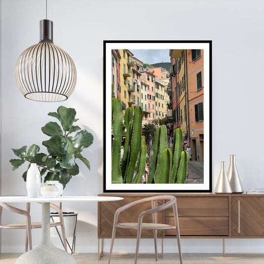 Plakat viser Riomaggiores hovedgate med sine fargerike hus. Sammenhengende gule og oransje hus ligger langsetter i byens eneste gate. En stor kaktus som vokser villig i dette klimaet. Her er motivet på plakat med hvit kant og i sort ramme.