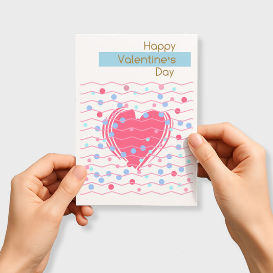 Kjærlighetskort med teksten Happy Valentine’s Day, stort rosa hjerte, grafiske linjer og prikker i pastellfarger. Person som holder kortet.