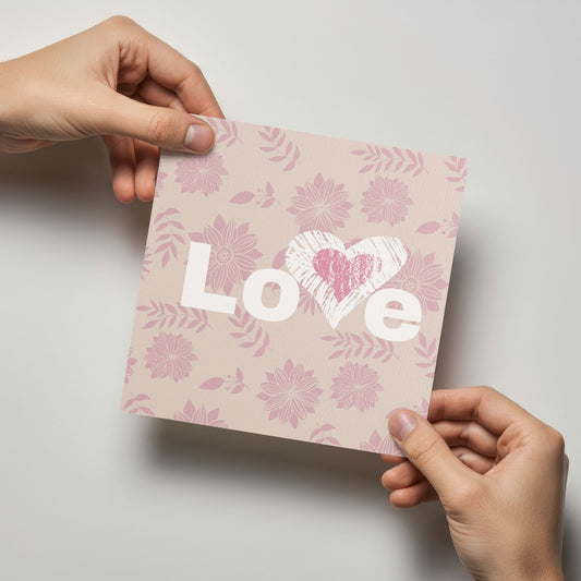 Valentineskort med teksten LOVE, dekorert med håndtegnet hjerte og rosa blomster i et mykt og romantisk design. Kortet blir holdt av en person.