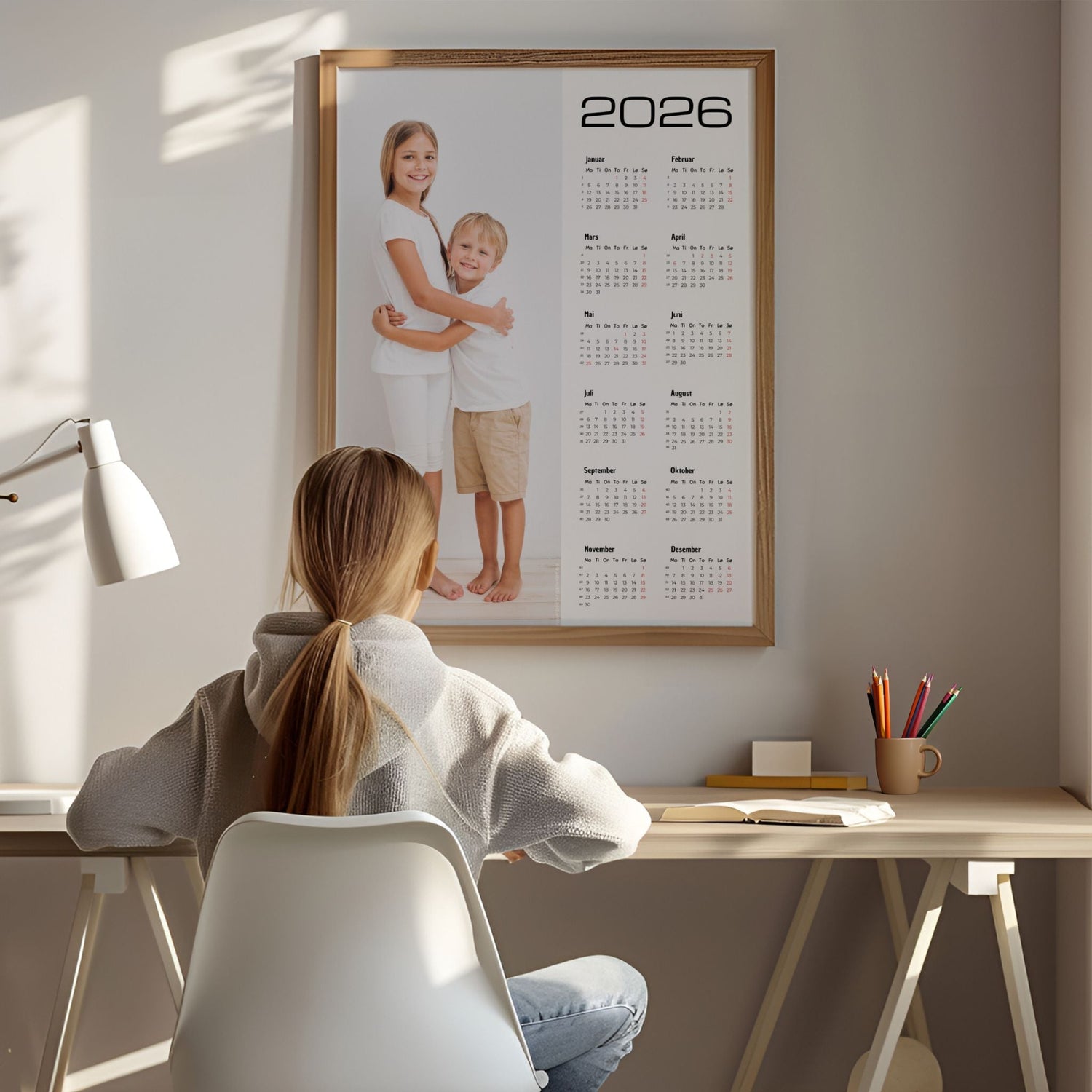 Personlig kalender 2026 i ramme med barnebilde, plassert over skrivebord i hjemmekontor.