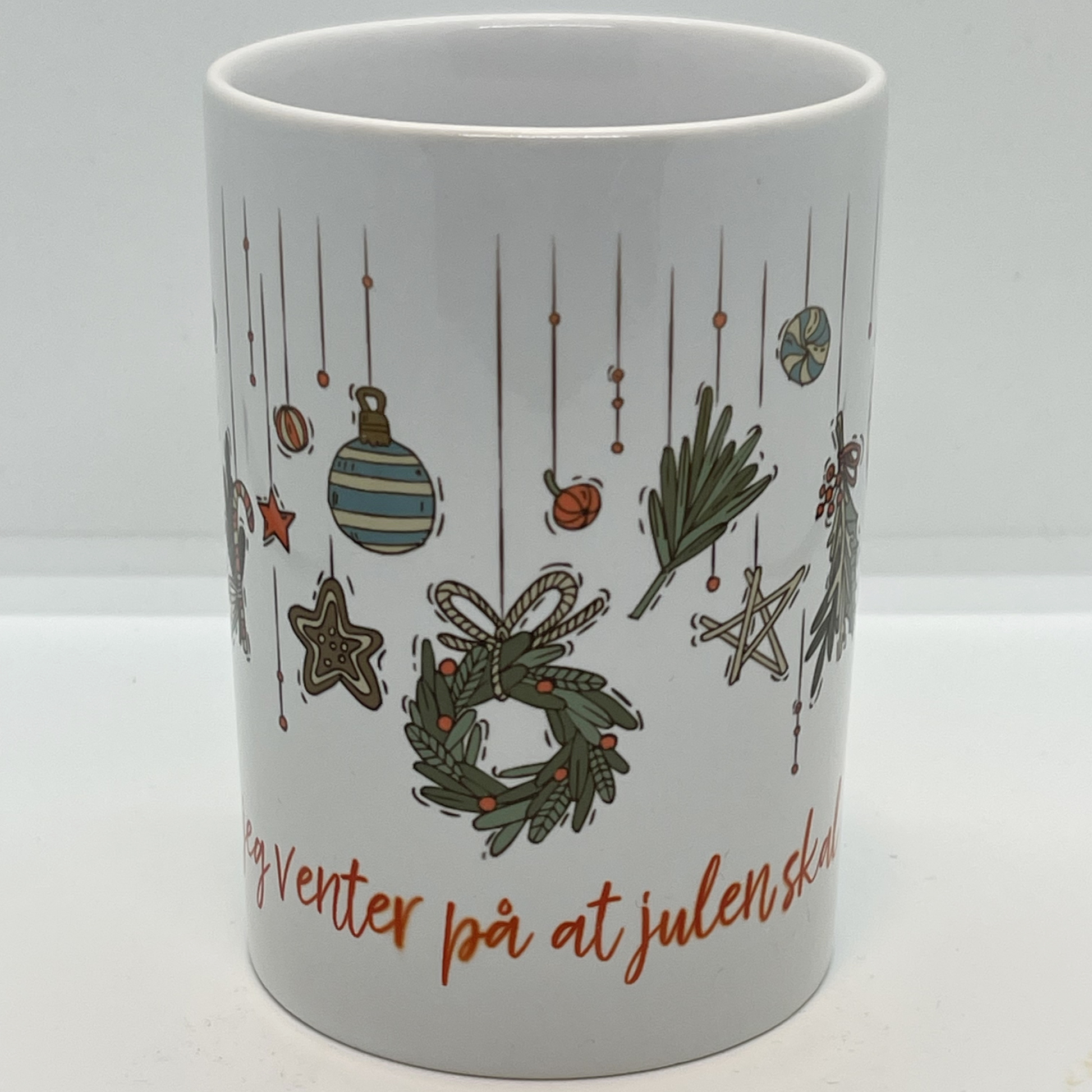 Hvitt krus i keramikk med grafisk julemotiv og håndskrevet rød tekst "-mens jeg venter på at julen skal komme". Volum: 29 cl. Diameter: 7 cm. Høyde: 10,5 cm.