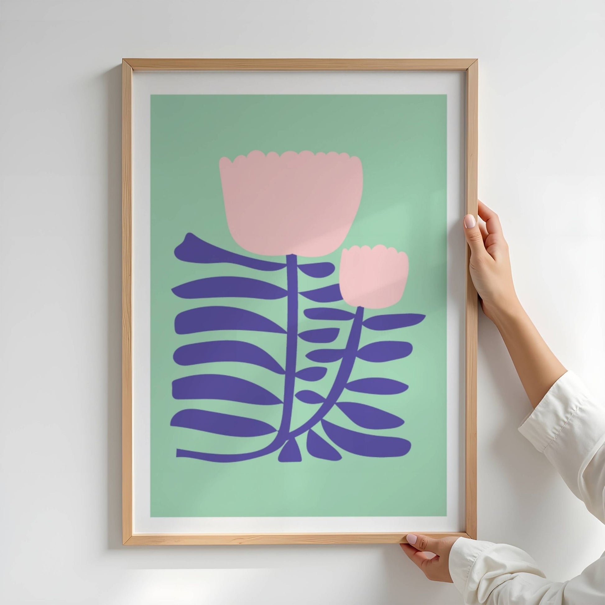 Bold Botanical No4 viser to lyserosa blomster strekker seg opp fra dype, blåfiolette blader i et stilisert og rytmisk uttrykk. Den mintgrønne bakgrunnen skaper en myk kontrast som løfter frem motivets delikate, men grafisk tydelige formspråk.  Illustrasjon viser plakat i eik ramme.