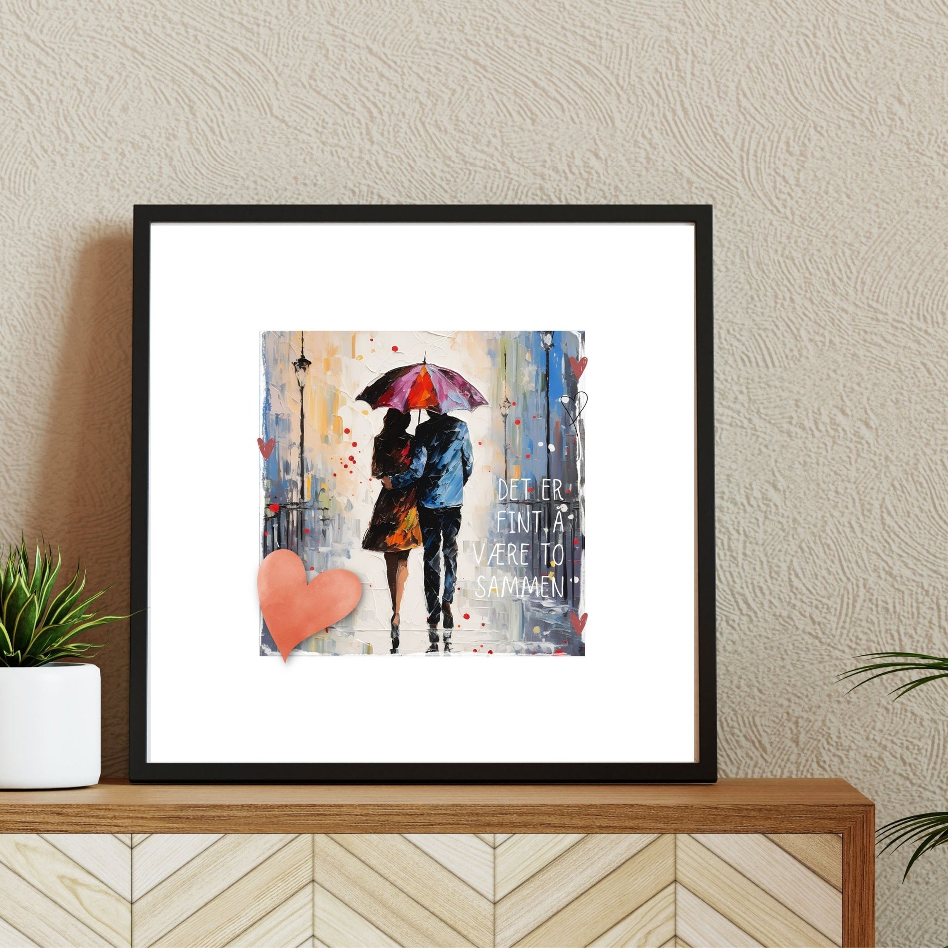 Plakat med rosa hjerte og tekst "Det er fint å være to sammen" - og et motiv med et kjærestepar som går under en rød paraply. Bildet har en hvit kant på 4 cm. Illustrasjon viser plakat i ramme.