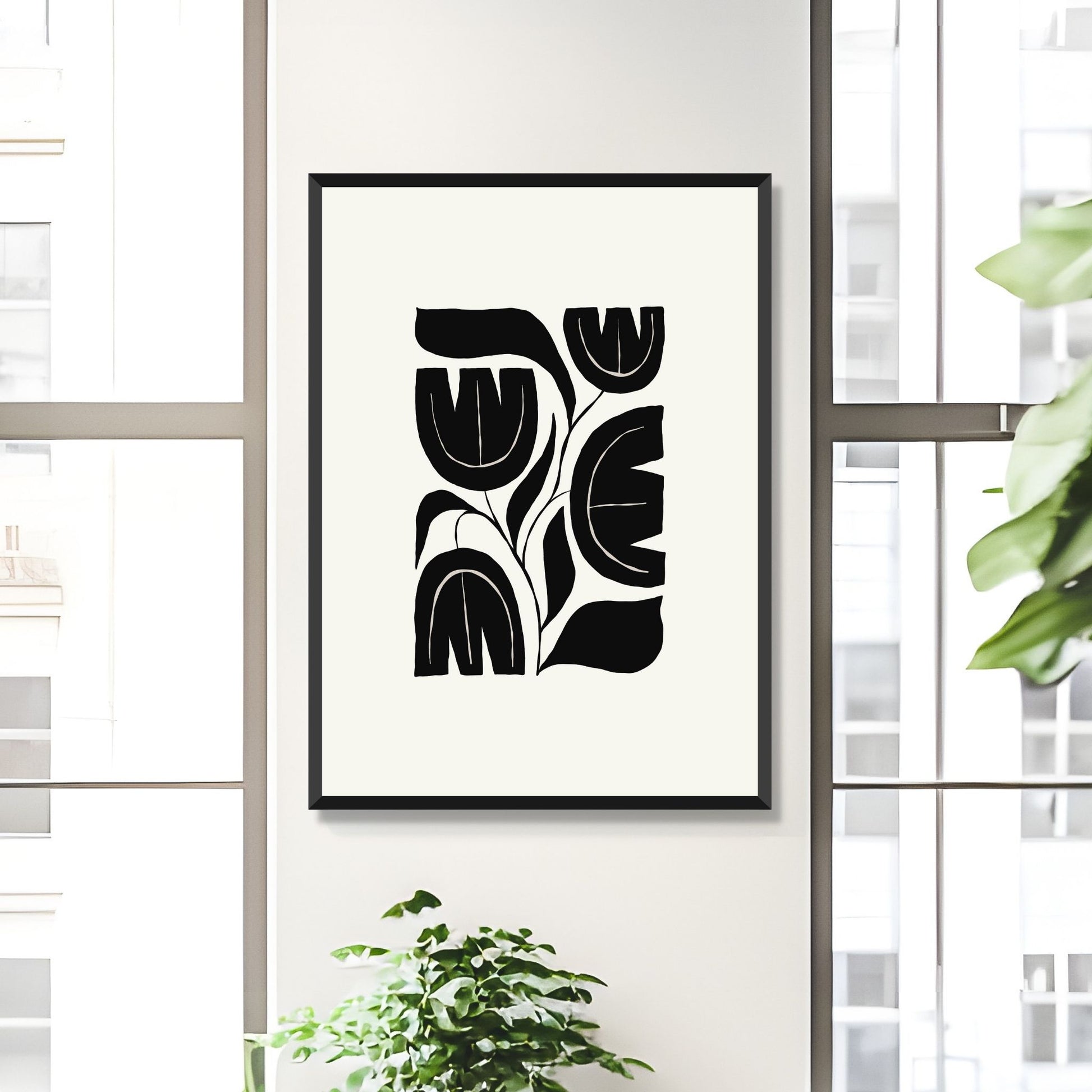Bildet viser en grafisk plakat i abstrakt, floralt mønster i sort og dempet hvit bakgrunn. Plakaten har en bredt kant i dempet hvit farge rundt og henger på en lys vegg mellom to vinduer i sort ramme.