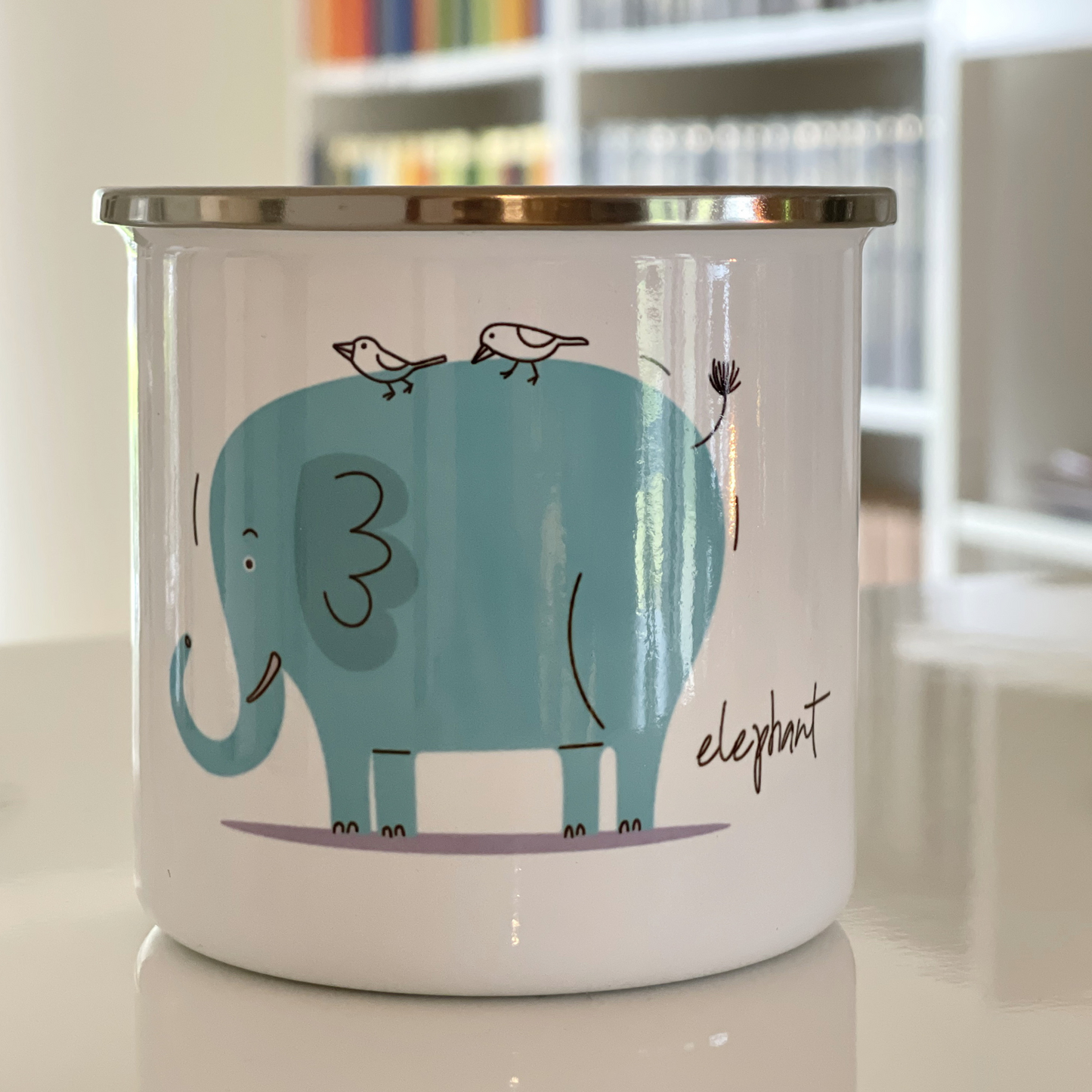 Emaljekrus i hvit farge med håndtak og en dekorativ sølvkant øverst. Kruset er påført et trykk med en grønn Elefant. Volum 32 cl. Diameter 8 cm.  Høyde 8 cm.