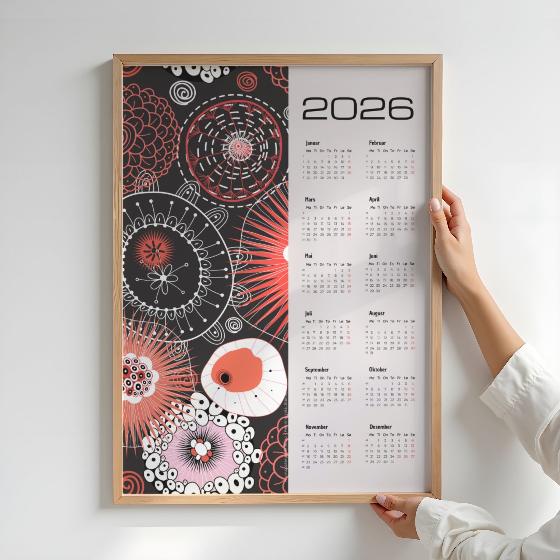 Grafisk kalender 2026 med røde og hvite abstrakte sirkler og linjemønstre på sort bakgrunn, med tydelig årskalender og norske helligdager. Illustrasjon viser kalender i eik ramme.