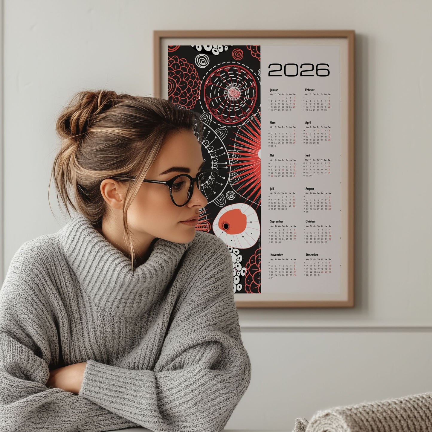 Grafisk kalender 2026 med røde og hvite abstrakte sirkler og linjemønstre på sort bakgrunn, med tydelig årskalender og norske helligdager. Illustrasjon viser kalender i eik ramme.