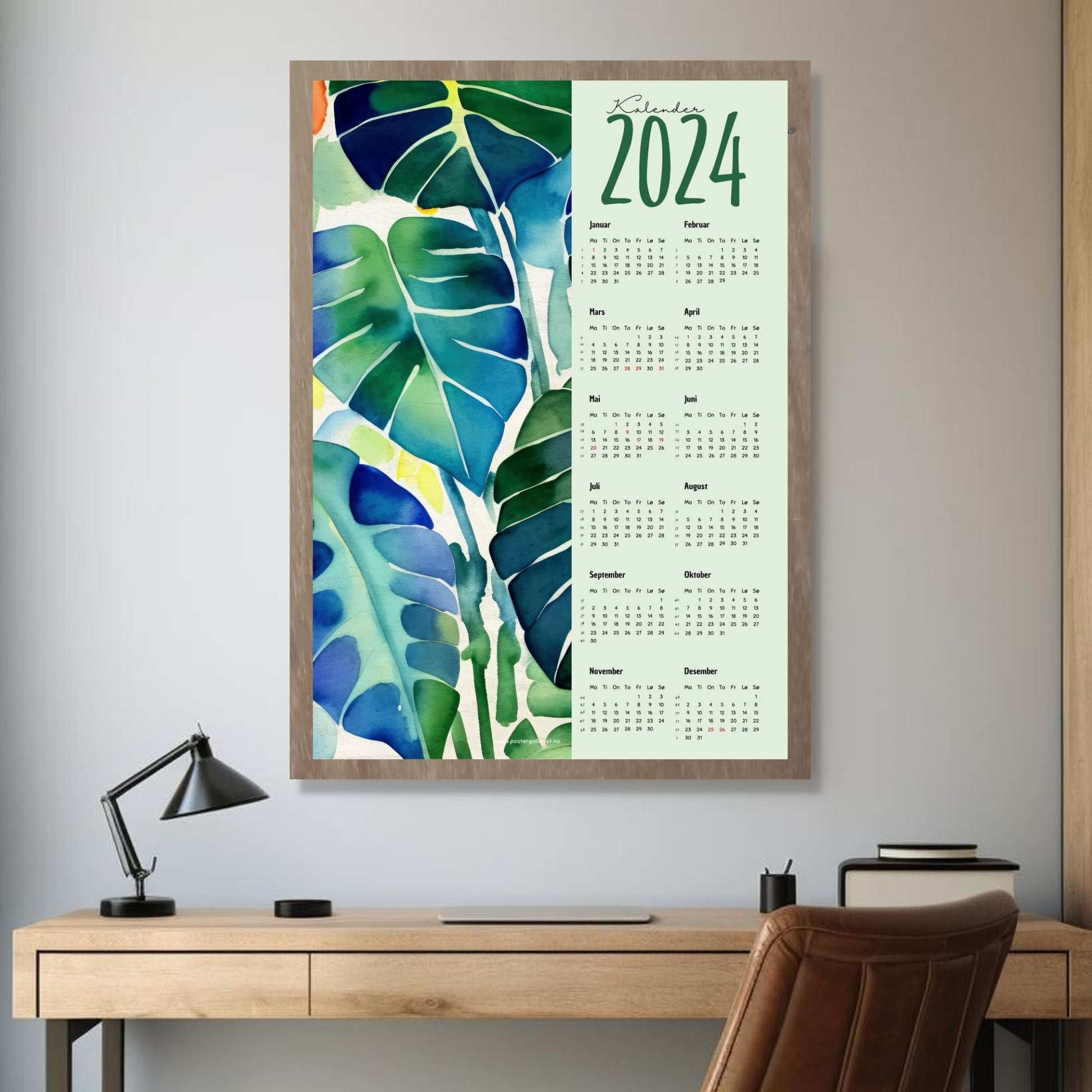 Kalender 2024 i grafisk print. Halve vertikale side har et grafisk, floralt motiv av tropiske blader i blå, grønne og turkise fargenyanser. Den andre halvdelen er påført 12 måneder med ukenummer og anmerket høytidsdager. Miljøbilde av kalender i eikeramme som henger på en vegg.