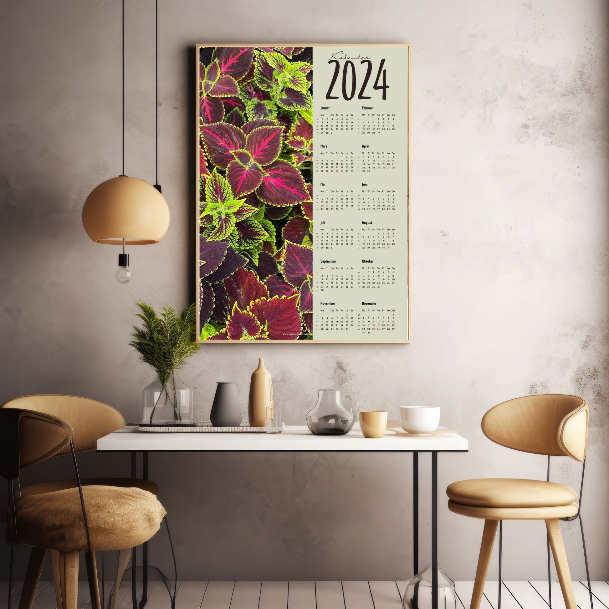 Kalender 2024 i grafisk print med fotomotiv. Halve vertikale side har et fotomotiv av en Coleus plante. Den andre halvdelen er påført 12 måneder med ukenummer og anmerket høytidsdager. Miljøbilde av kalender i eikeramme som henger på en vegg.