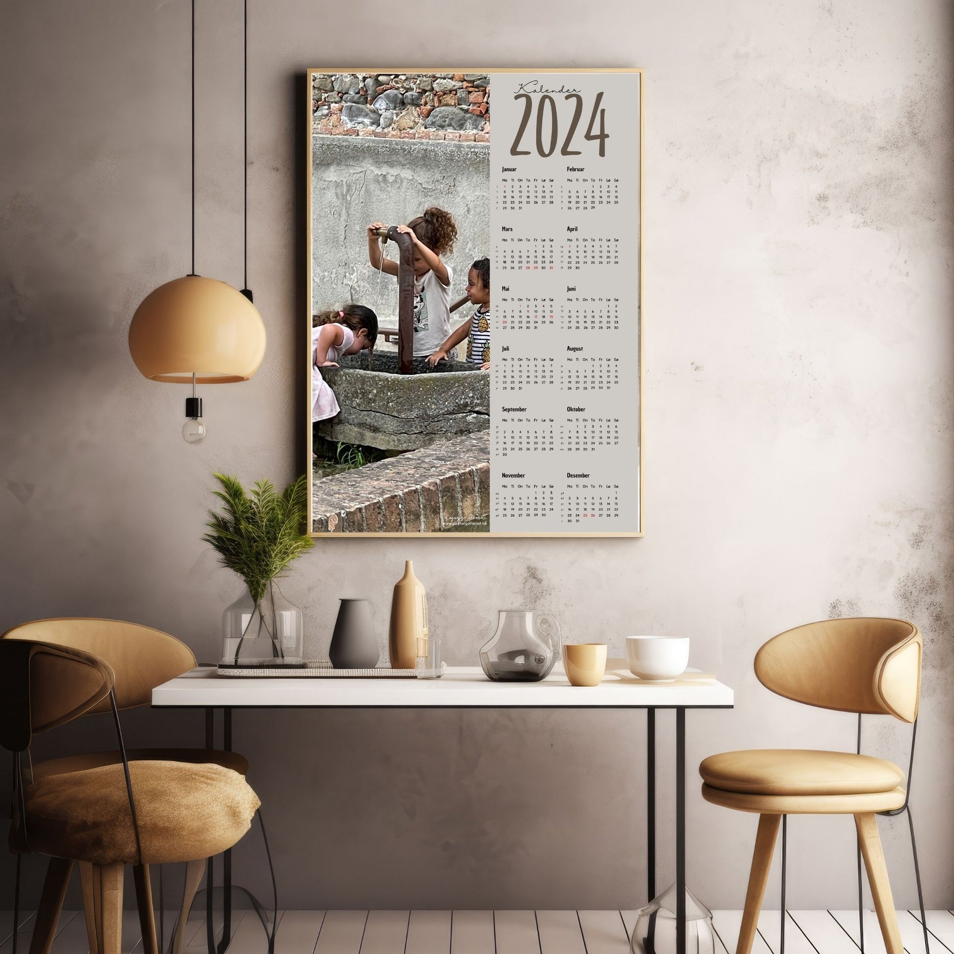 Kalender 2024 i grafisk print med fotomotiv. Halve vertikale side er et fotografi av noen småjenter som avkjøler seg ved en vannfontene i Grinzane, Italia. Den andre halvdelen er påført 12 måneder med ukenummer og anmerket høytidsdager. Miljøbilde av kalender i eikeramme som henger på en vegg.