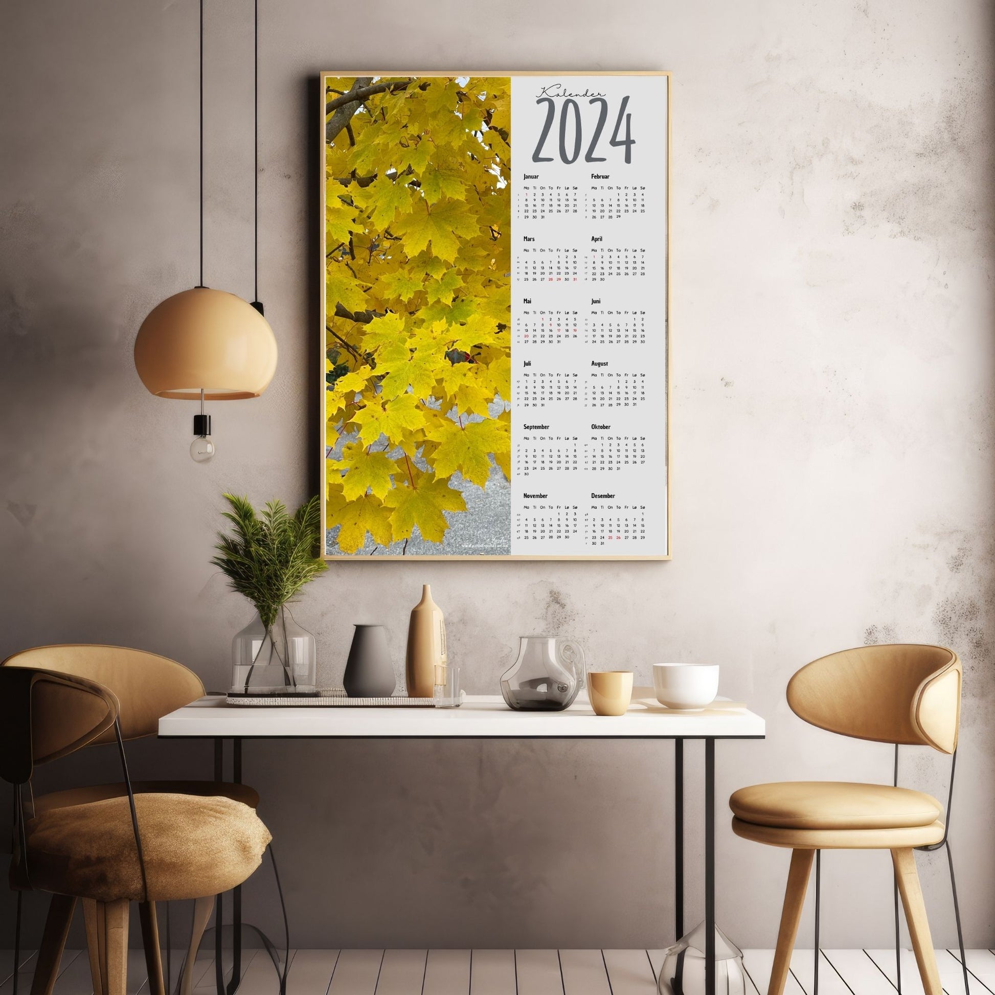 Kalender 2024 i grafisk print med fotomotiv. Halve vertikale side er et fotografi en Lønn i høstgule farger. Den andre halvdelen er påført 12 måneder med ukenummer og anmerket høytidsdager. Miljøbilde av kalender i eikeramme som henger på en vegg.