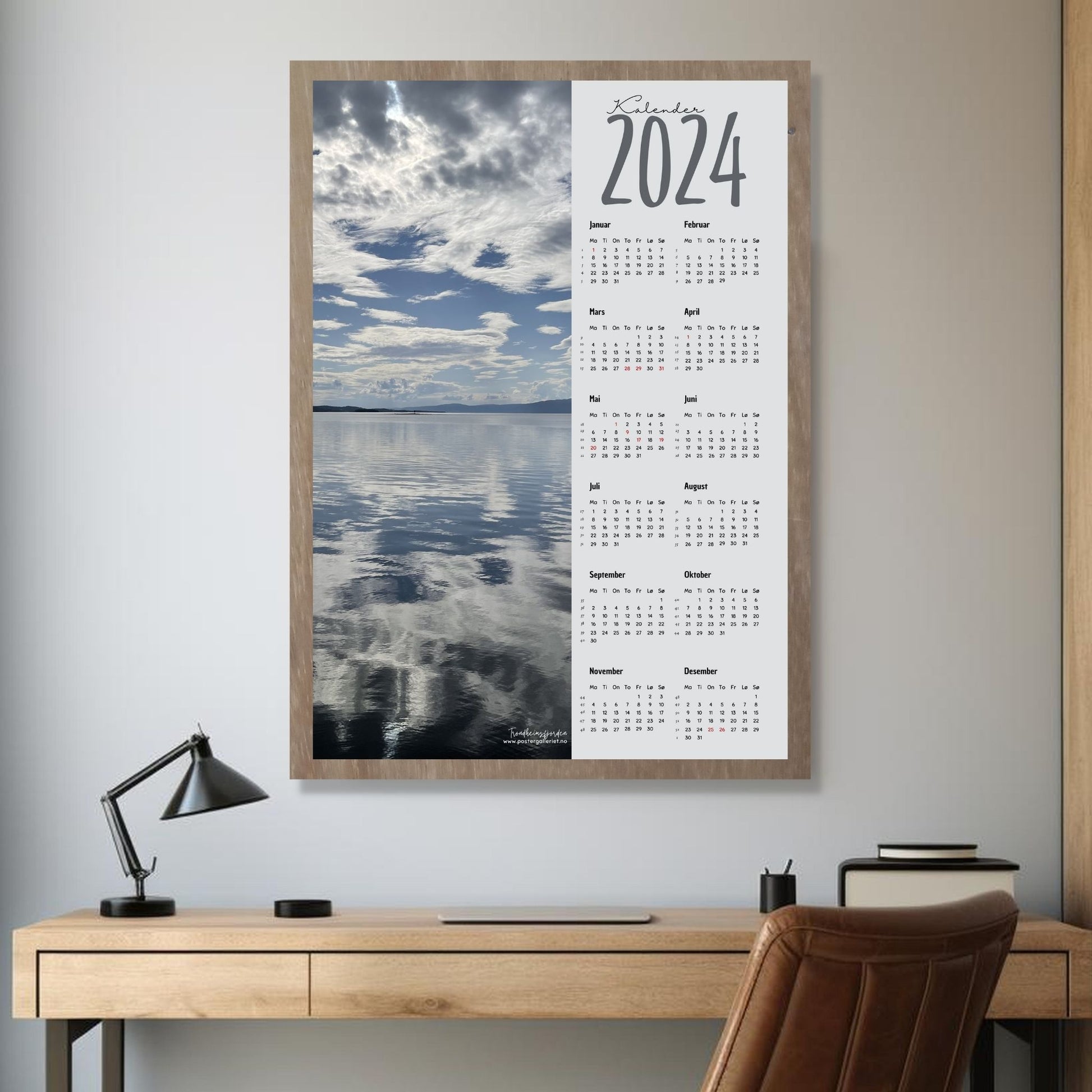 Kalender 2024 i grafisk print med fotomotiv. Halve vertikale side er et fotografi fra en vakker sommerdag ved Trondheimsfjorden ved Steinkjer. Den andre halvdelen er påført 12 måneder med ukenummer og anmerket høytidsdager. Miljøbilde av kalender i eikeramme som henger på en vegg.