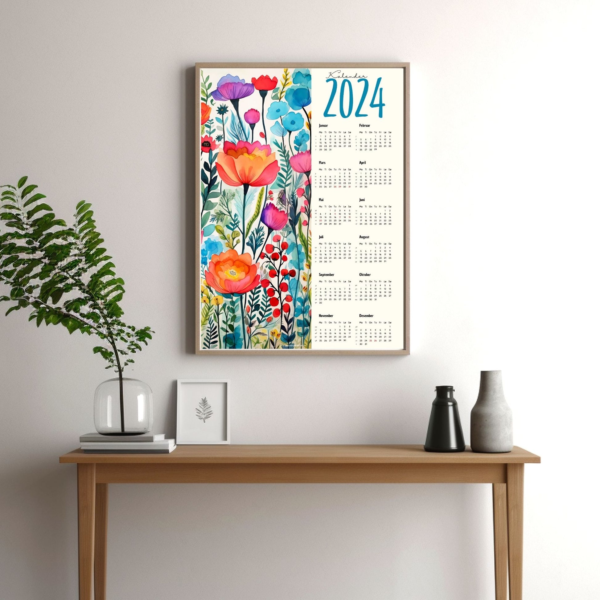 Kalender 2024 i grafisk print. Halve vertikale side har et grafisk floralt motiv i friske fargenyanser. Den andre halvdelen er påført 12 måneder med ukenummer og anmerket høytidsdager. Miljøbilde av kalender i eikeramme som henger på en vegg.