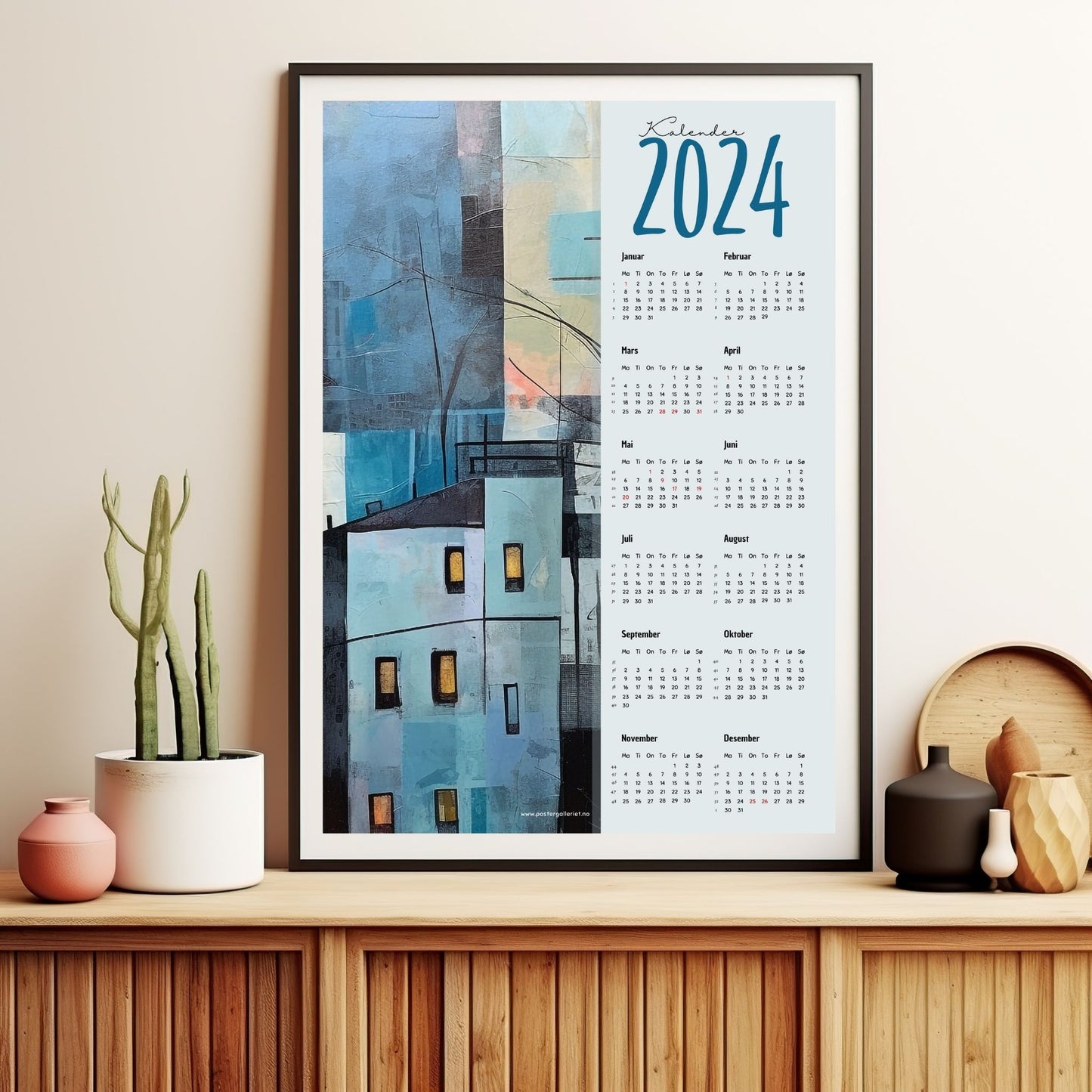 Kalender 2024 i grafisk print. Halve vertikale side har et grafisk motiv av en by i blåe fargenyanser med innslag av beige. Den andre halvdelen er påført 12 måneder med ukenummer og anmerket høytidsdager. Miljøbilde av kalender i en sort ramme som står på et skap.