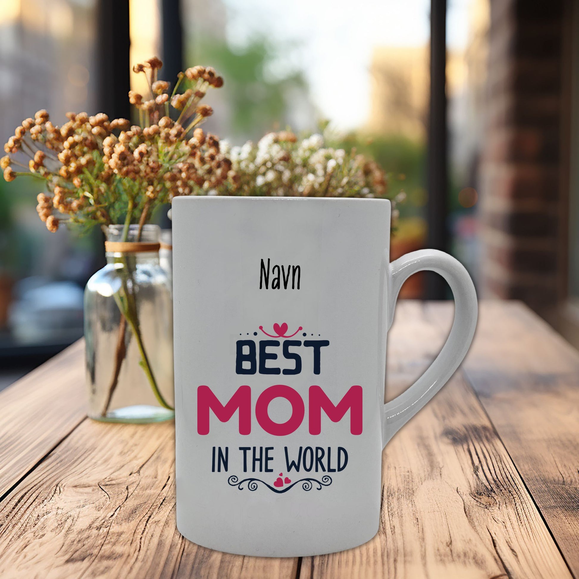 Krus i hvit keramikk med påskrift "Best mom in the world" på en side, og tre hjerter på motsatt side. Kruset kan personaliseres med navn. Trykk i sort og rosa.