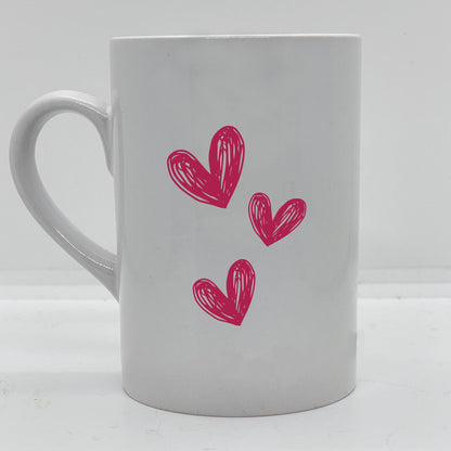 Krus i hvit keramikk med påskrift "Best mom in the world" på en side, og tre hjerter på motsatt side. Trykk i sort og rosa.