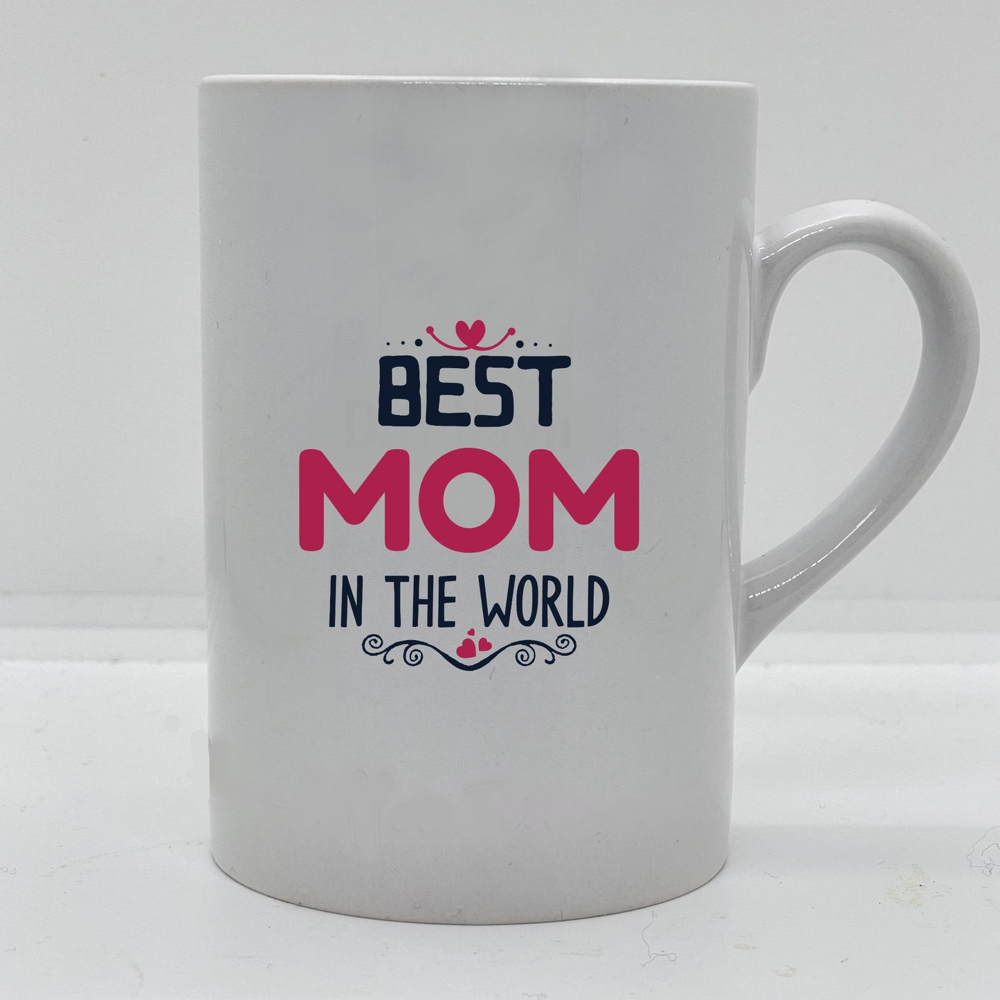Krus i hvit keramikk med påskrift "Best mom in the world" på en side, og tre hjerter på motsatt side. Trykk i sort og rosa.