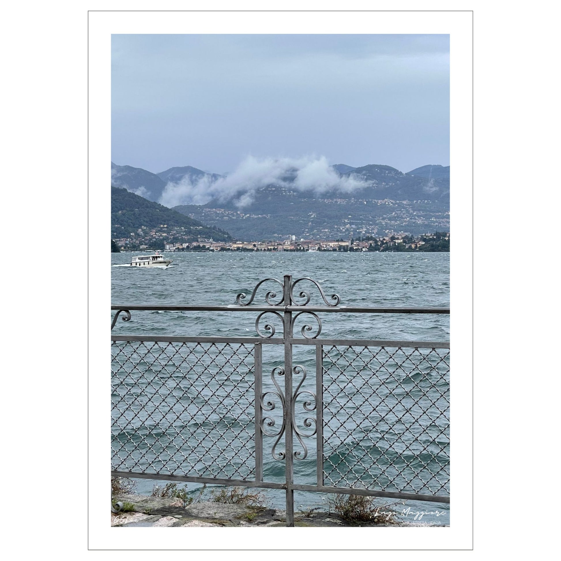 En regntung og vindfull augustdag i Stresa ved Lago Magiore, Piemonte. Det er lavt skydekke og bølger på sjøen. Et gjerde i smijern skiller parken og sjøen. I bakkant skimtes Alpene og byen Verbania.  