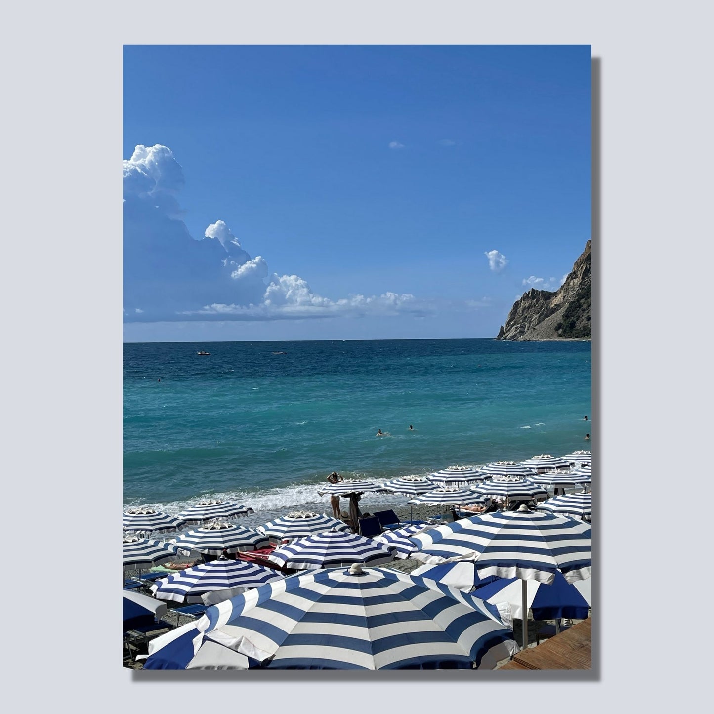 Motiv fra Montereosso al Mare, en av de fem byene i Cinque Terre ved Liguriakysten i Italia. Bildet viser blått middelhav og himmel som helt ned i vannet i horisonten. På høyre side skimtes noen klipper. På sranden er det tett i tett med blå og hvitstripete parasoller. Bildet vise mockup bilde av motiv på lerret.