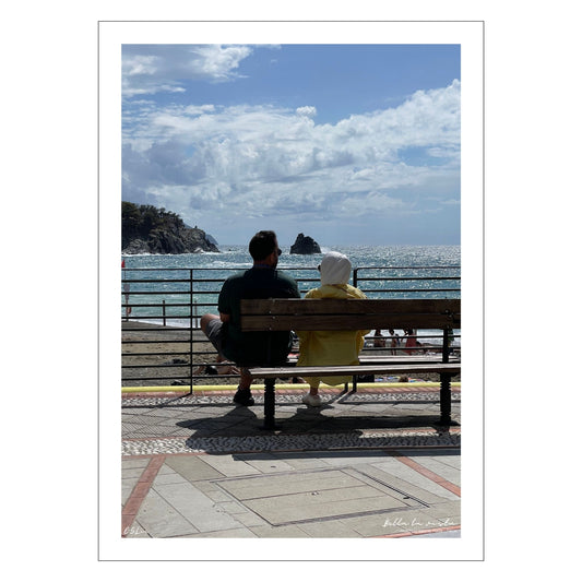 Motiv fra Montereosso al Mare, en av de fem byene i Cinque Terre ved Liguriakysten i Italia. Bildet viser blått middelhav og himmel som helt ned i vannet i horisonten. Her har et par funnet seg godt til rette på en av byens benker. Fri sikt utover Middelhavet så langt øyet ser. Til venstre på bildet går klipper ned i havet. Blå himmel er dekket med noen hvite skyer. Hav og himmel møtes ute i horisonten. 