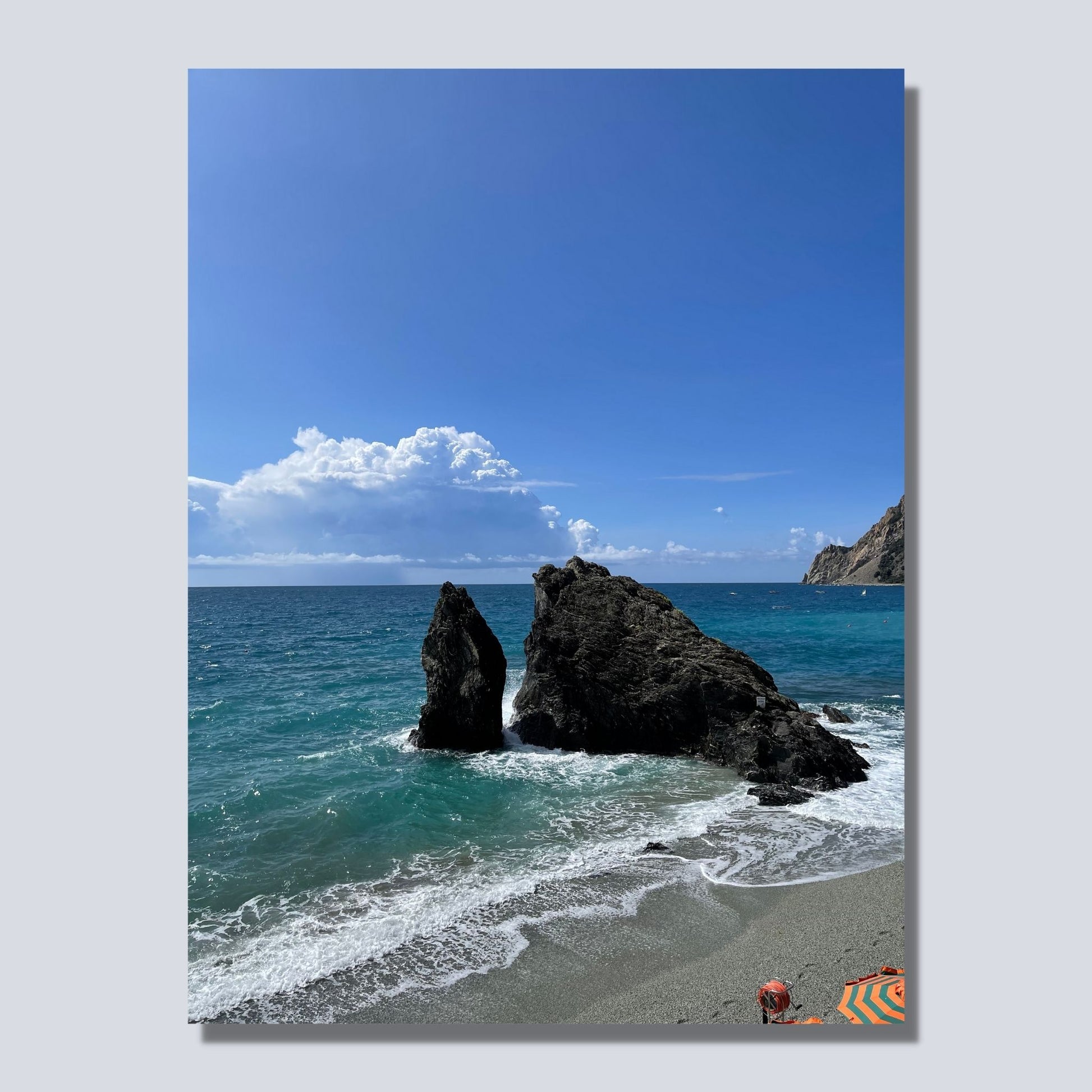 Motiv fra stranden ved Montereosso al Mare, en av de fem byene i Cinque Terre ved Liguriakysten i Italia. Bildet viser blått middelhav og himmel som møter havet i horisonten. . I overgangen mellom himmel og hav ligger en stor skybanke. Det slår små bølger inn mot den karakteristiske klippen som ligger midt på stranden . På dette bildet presenteres motivet  som lerret.