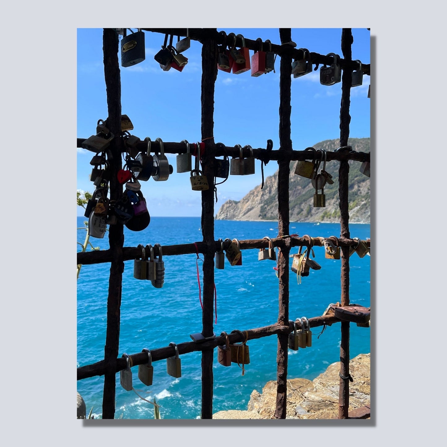 Bildet viser en mur med en åpning med et gitter, der det er hengt opp hengelås med håp om et langt liv i kjærlighet. Gjennom gitteret ses det blåe Middelhavet og klippene som går ned i sjøen. Bildet er på lerret.