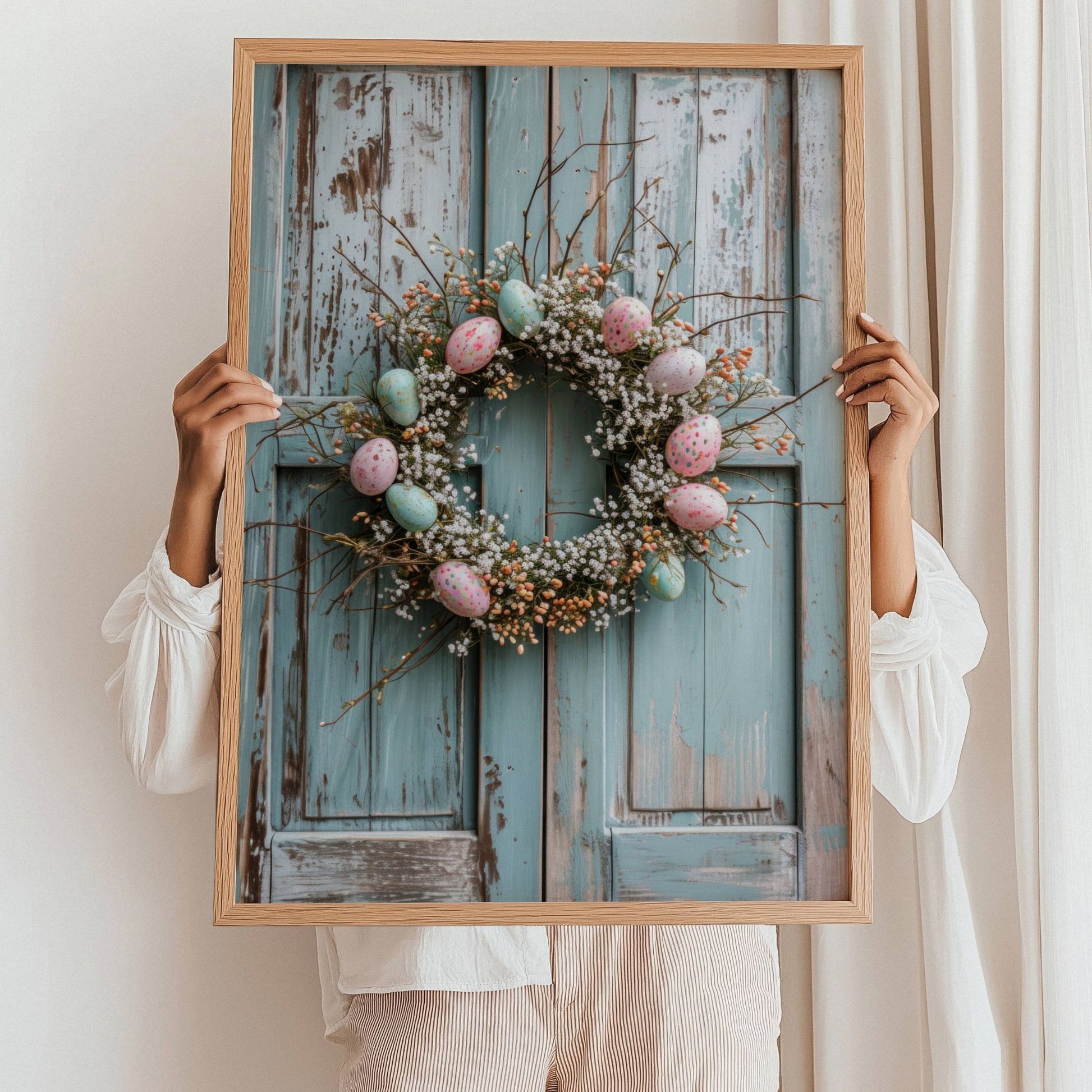 Grafisk illustrasjon av en krans av kvister og blomster, pyntet med delikate påskeegg i duse pastellfarger. Kransen henger vakkert på en gammel, rustikk dør. Illustrasjonsfoto viser plakat i sort ramme som står i gulvet lent mot en vegg.