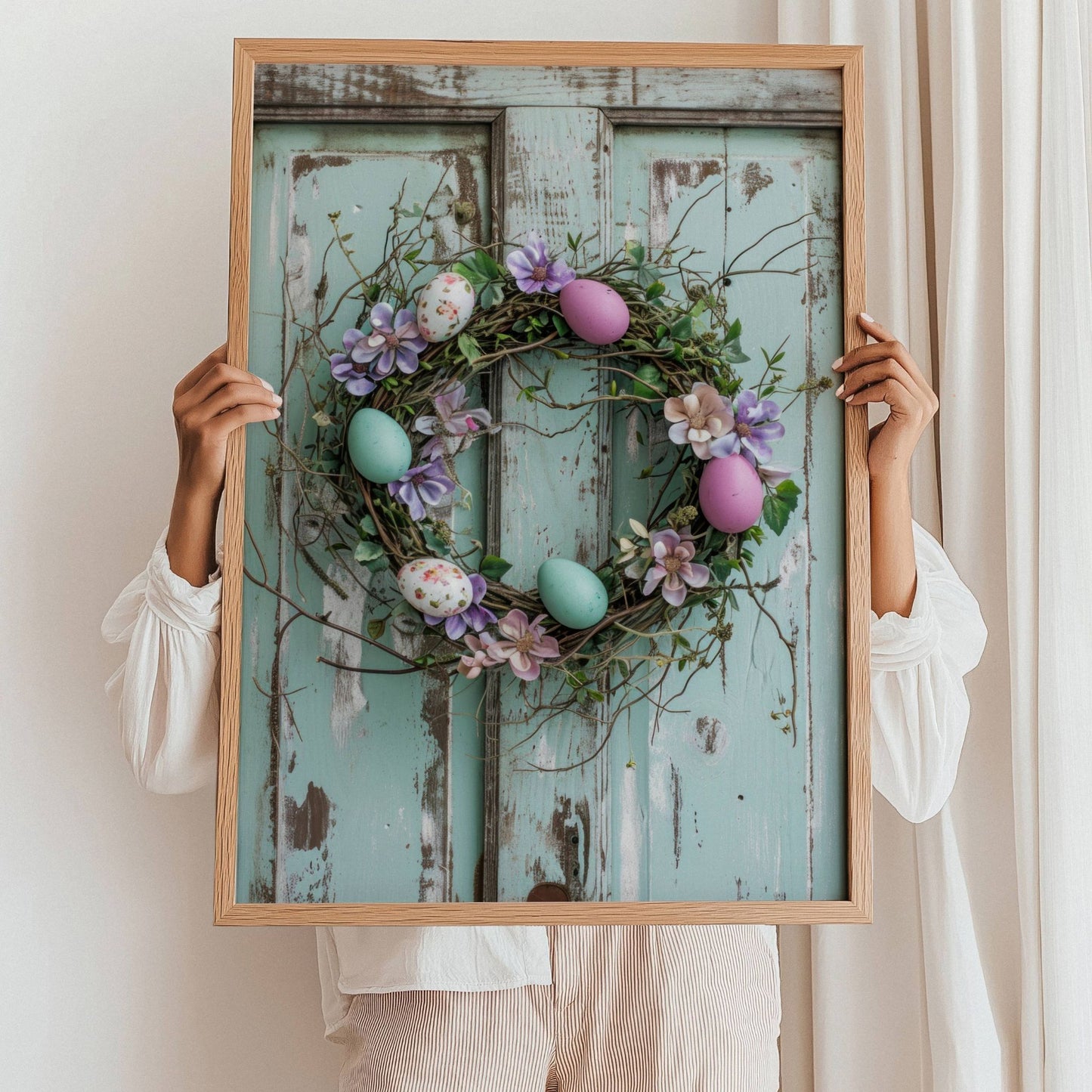 Grafisk illustrasjon av en krans av kvister og blomster, pyntet med delikate påskeegg i duse pastellfarger. Kransen henger vakkert på en gammel, rustikk dør. Illustrasjonsfoto viser plakat i sort ramme som står i gulvet lent mot en vegg.