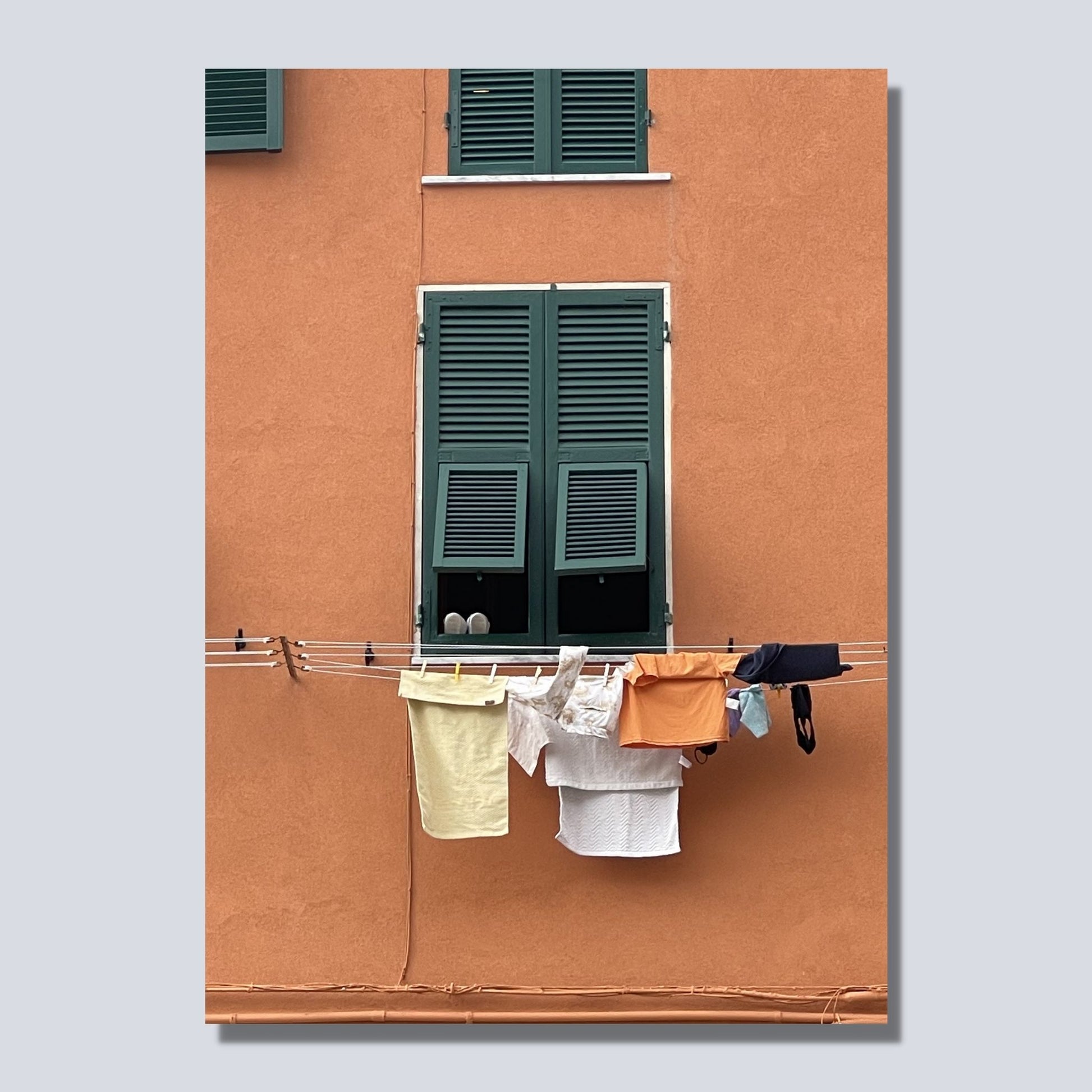 Plakat viser motiv er fra Riomaggiore, en av de fem byene i Cinque Terre ved Liguriakysten i Italia. Riomaggiore ligger lengst østover av de 5 byene. Her er det som i alle Italienske byer, klestørk på kryss og tvers på alle husfasader. På plakaten er det en oransje fasadevegg med et vindu med grønne skodder. Under vinduet henger dagens klesvask til tørk.  Bilde av motiv på lerret.