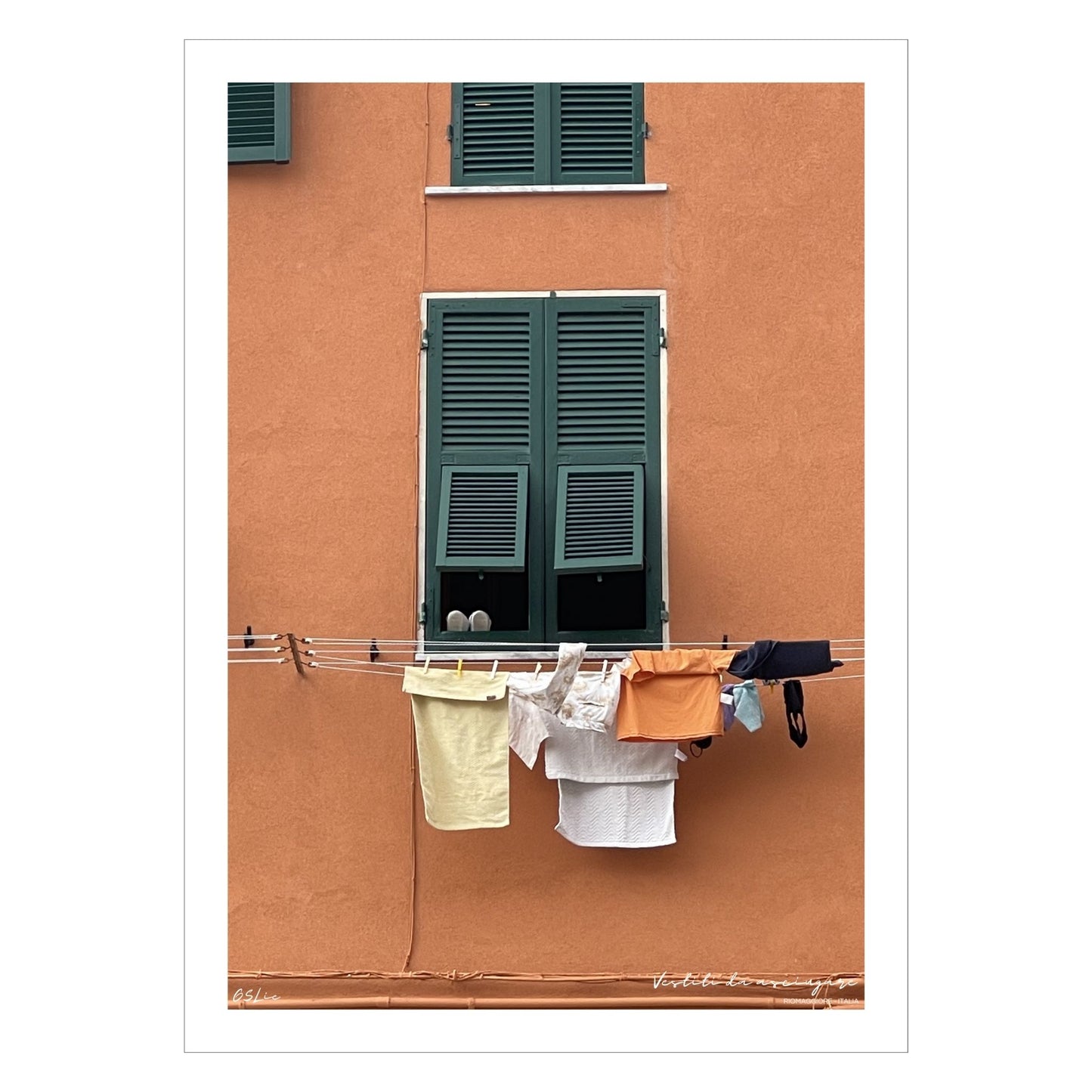 Plakat viser motiv fra Riomaggiore, en av de fem byene i Cinque Terre ved Liguriakysten i Italia. Riomaggiore ligger lengst østover av de 5 byene. Her er det som i alle Italienske byer, klestørk på kryss og tvers på alle husfasader. På plakaten er det en oransje fasadevegg med et vindu med grønne skodder. Under vinduet henger dagens klesvask til tørk.  